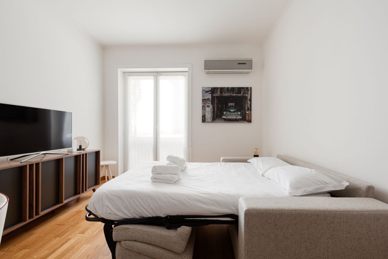 A modern hotel room with a bed, a TV, a desk, and a window.
