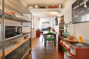 A kitchen with a refrigerator, shelves with dishes, a dining table with chairs, and a view of the outside.