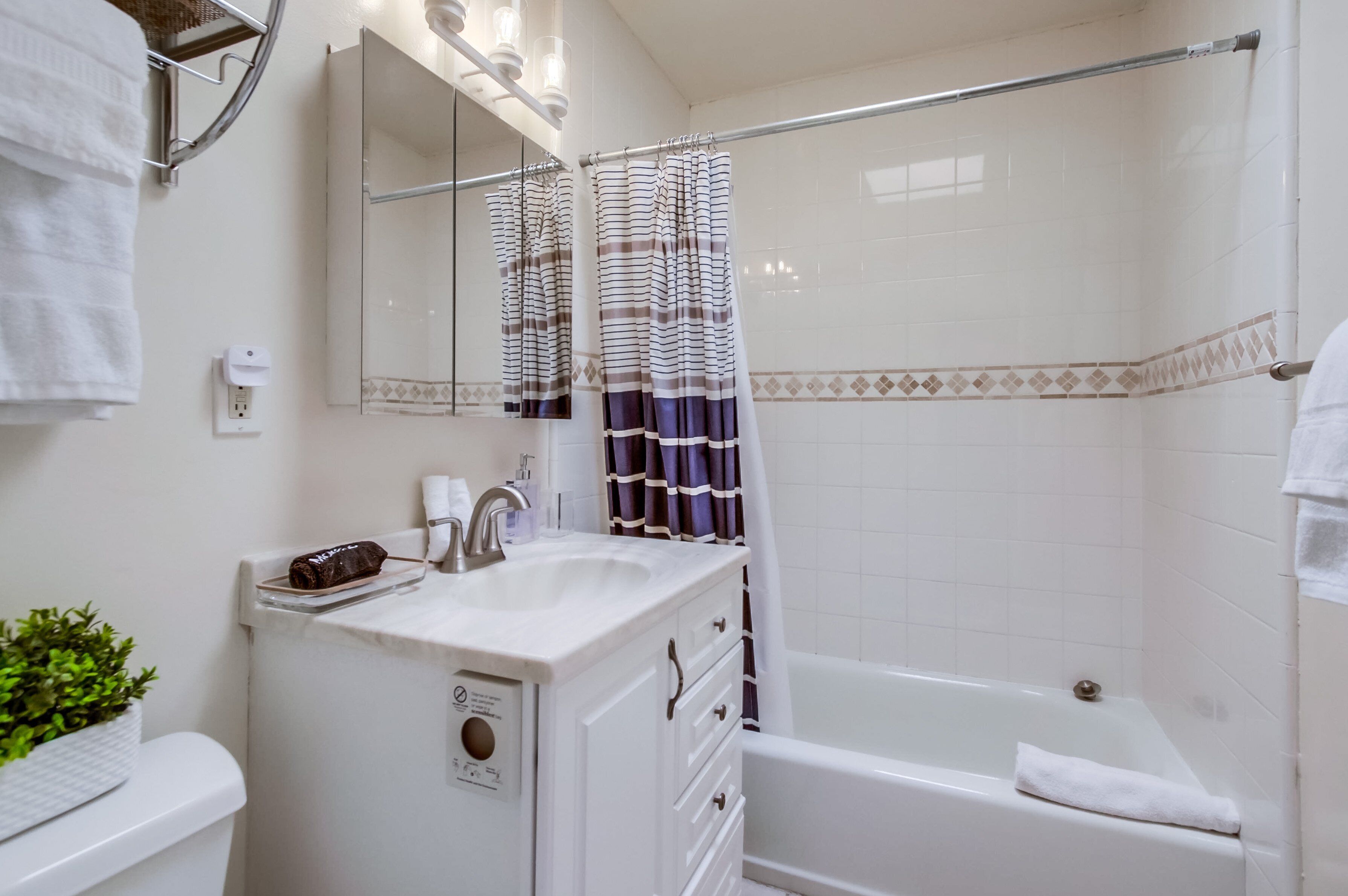 Ein Badezimmer mit einem weißen Waschbecken, einem Spiegel und einer Badewanne. Über dem Waschbecken befindet sich ein Handtuchhalter und ein Duschvorhang mit horizontalen Streifen.