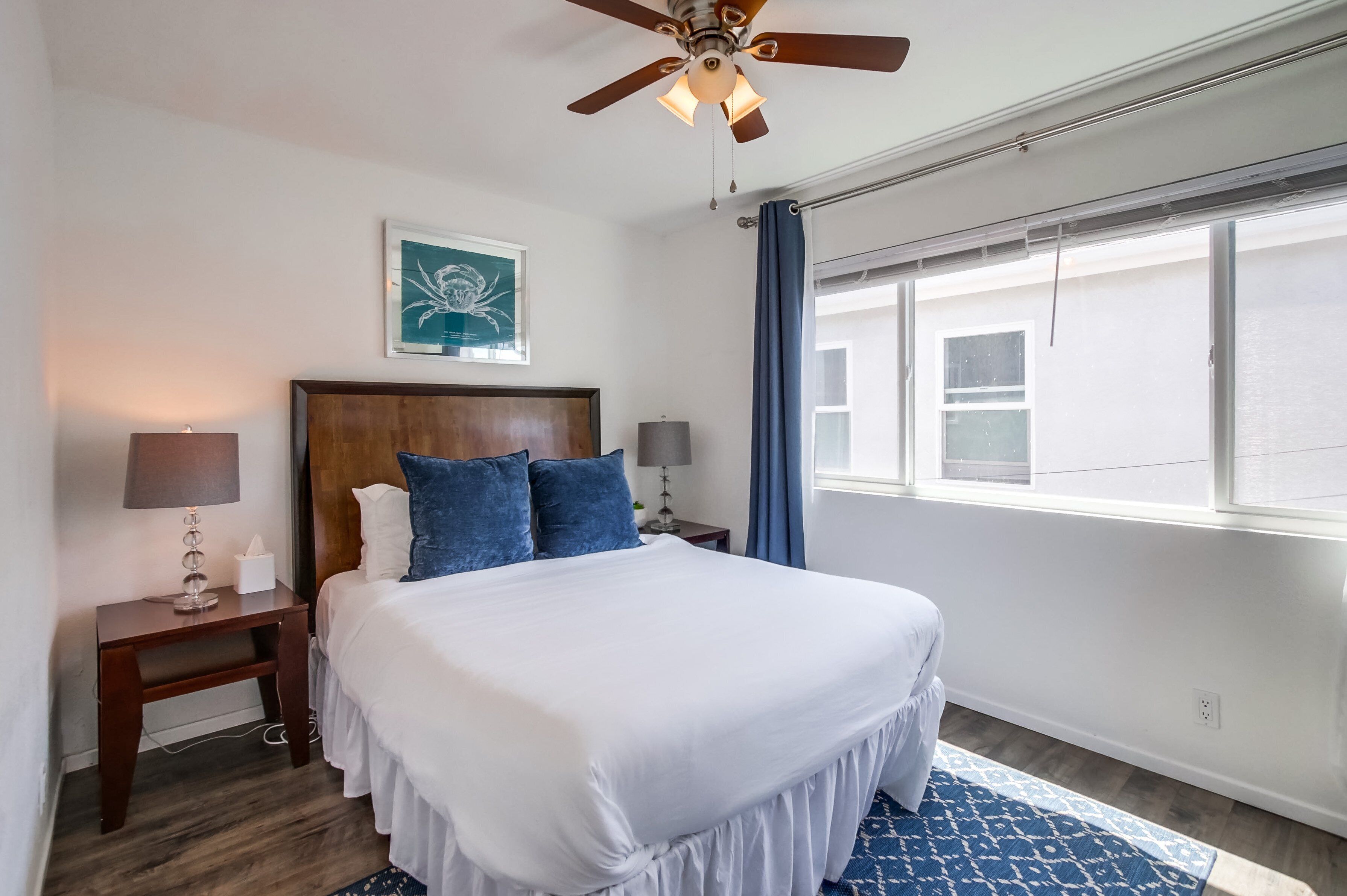 A hotel room with a large bed, two bedside lamps, a ceiling fan, a window with curtains, and a framed artwork on the wall.