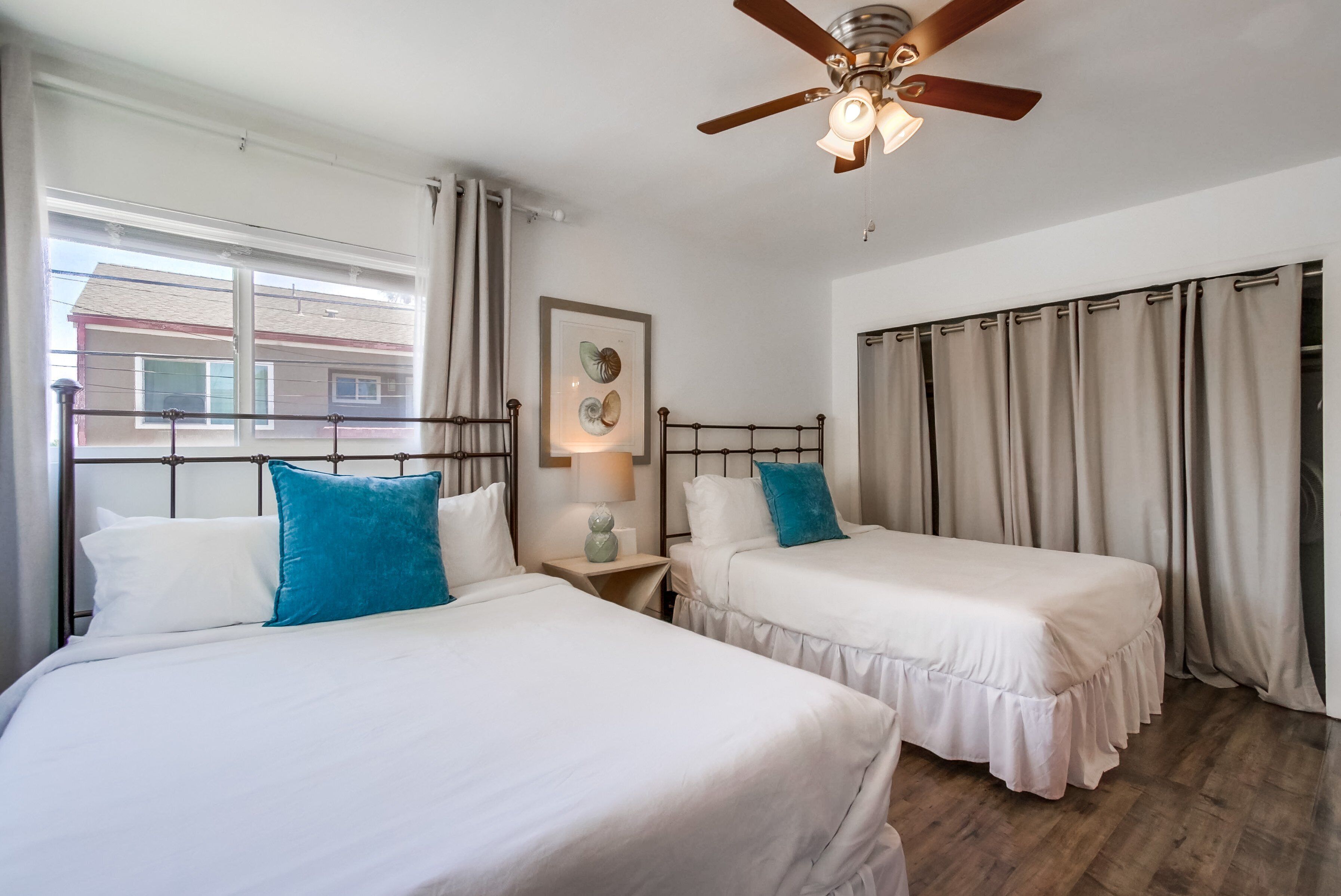 Un dormitorio con dos camas, un ventilador de techo, una ventana con cortinas y un cuadro en la pared.