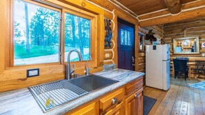 A log cabin interior with a kitchen, dining area, and a view of the forest through large windows. - Pine Cabin- Log cabin close to Custer (Custer)
