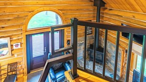 A log cabin interior with a wooden staircase, a stone fireplace, and a view through a window. - Pine Cabin- Log cabin close to Custer (Custer)
