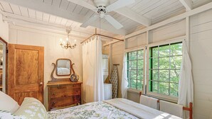 Une chambre avec une porte en bois, une commode, un lit avec une literie à motifs floraux, un ventilateur de plafond et une fenêtre avec des rideaux.