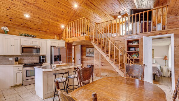 A spacious kitchen with a central island, wooden staircase, and a dining area with a large table and chairs. - Greers Ferry Home w/ Fire Pit Near Shiloh Marina! (Greers Ferry)