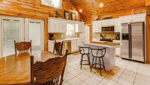 A wooden-paneled kitchen with white cabinets, a stainless steel refrigerator, and a dining area with a wooden table and chairs. - Greers Ferry Home w/ Fire Pit Near Shiloh Marina! (Greers Ferry)