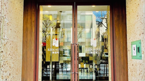 Entrada de un hotel con puerta de vidrio, marco de madera y toldo rojo.