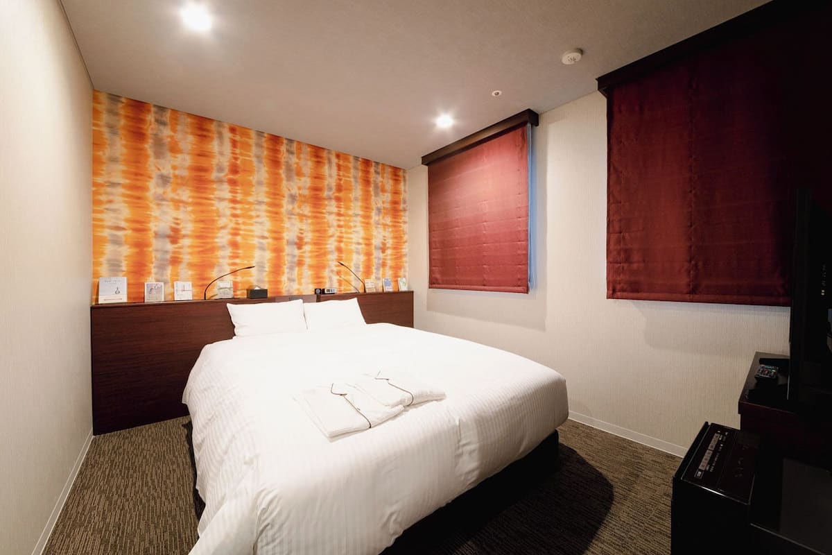 A hotel room with a large bed, a headboard, a wall-mounted TV, and a window with red curtains.