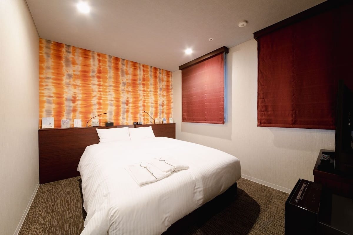 A hotel room with a large bed, a headboard, a wall-mounted TV, and a window with red curtains.