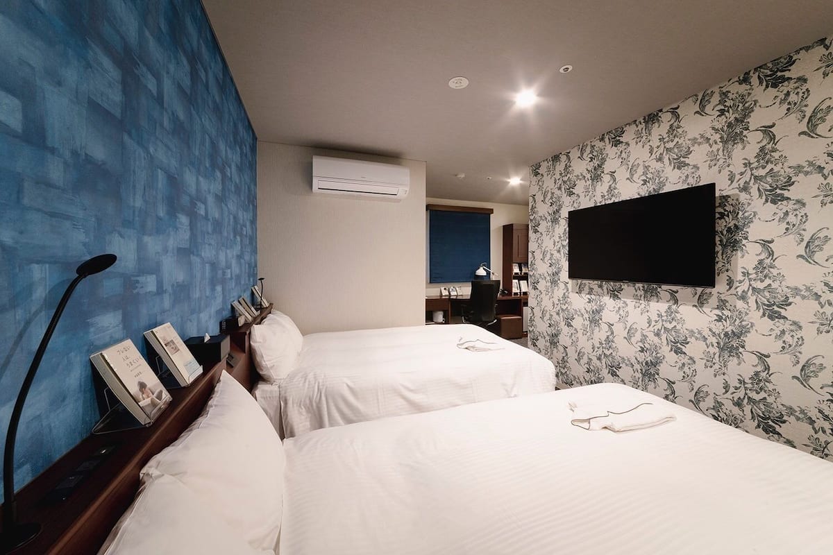 A hotel room with two beds, a flat-screen TV mounted on the wall, a desk with a chair, and a blue accent wall.