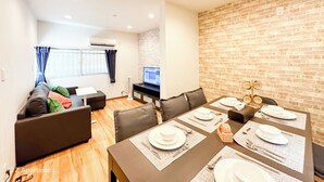 A modern living room with a dining area set for four, featuring a brick wall, a flat-screen TV, and a sofa with cushions.