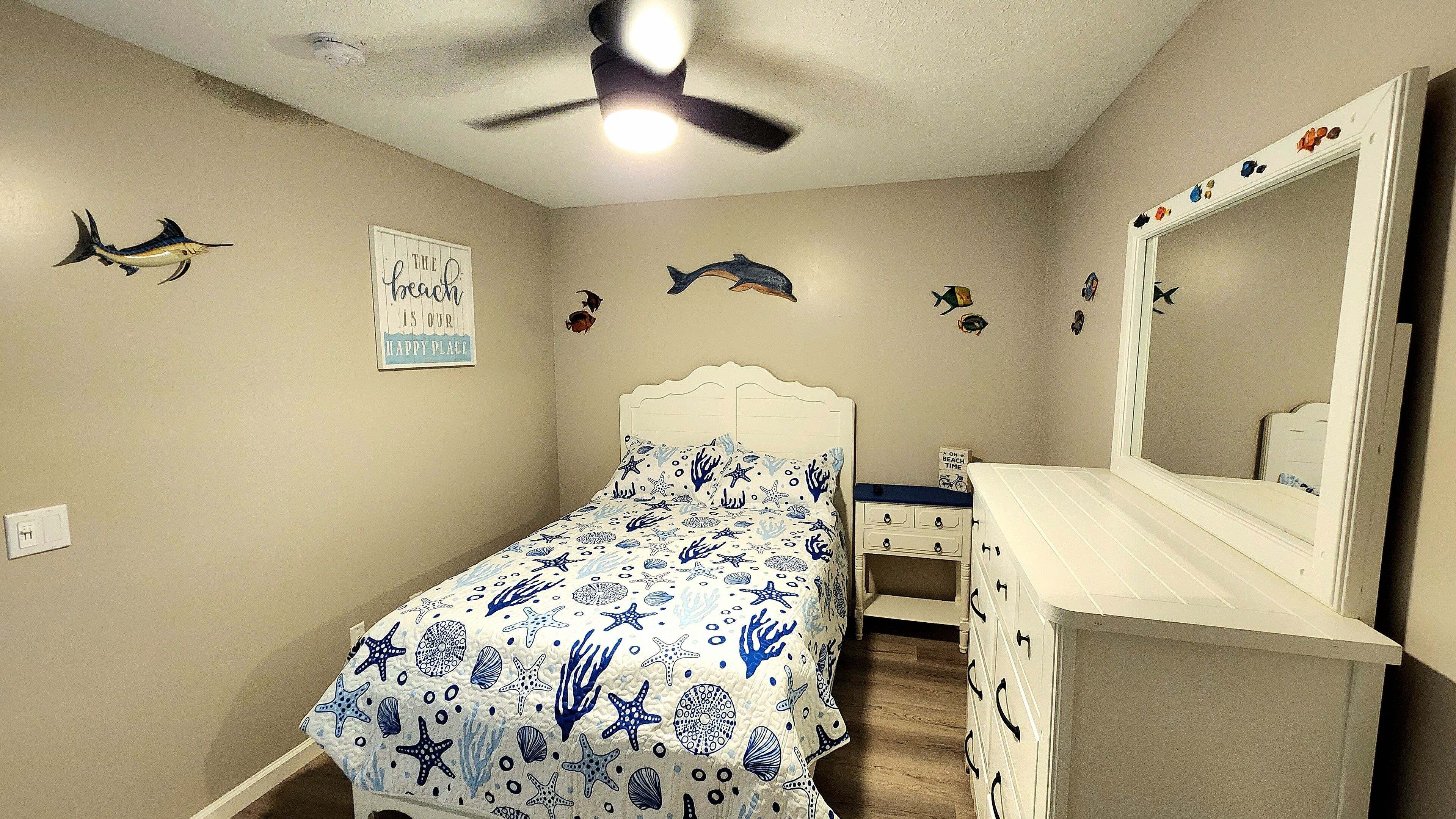 Een slaapkamer met een bed, een dressoir, een spiegel en wanddecoraties, waaronder vissen en een bord met de tekst 'De oceaan is onze gelukkige plek'.