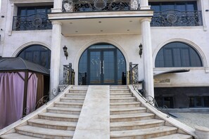 Une entrée imposante, avec un escalier en marbre menant à une grande porte en arc de cercle, flanquée de balustrades en métal ouvragé et surmontée d’un auvent.