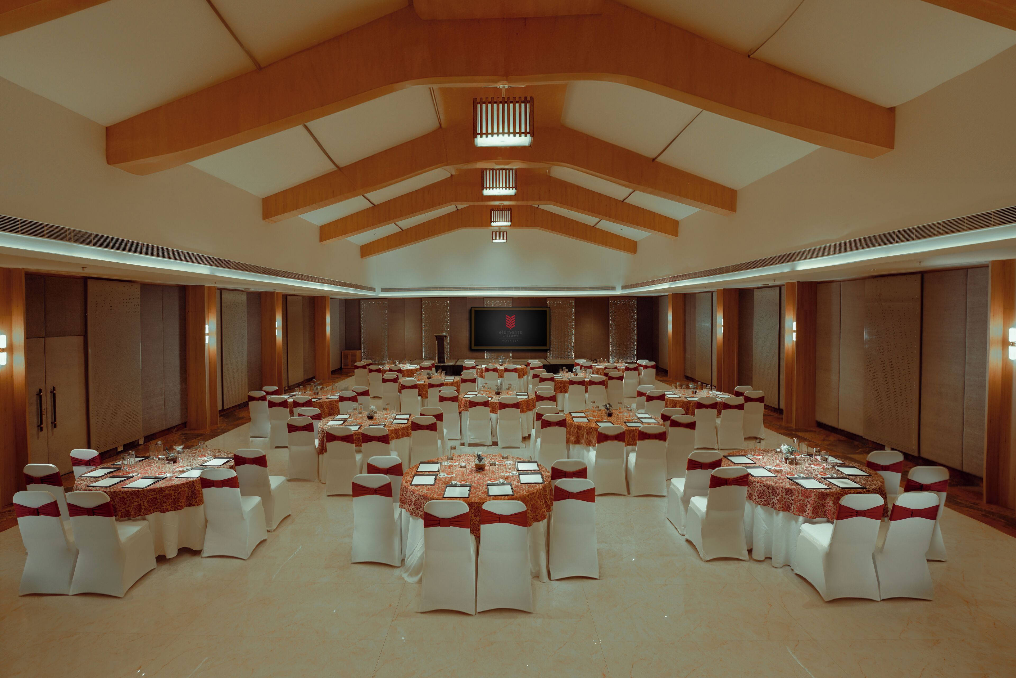 Un amplio salón de banquetes con múltiples mesas redondas, cada una dispuesta con sillas blancas y cintas rojas.