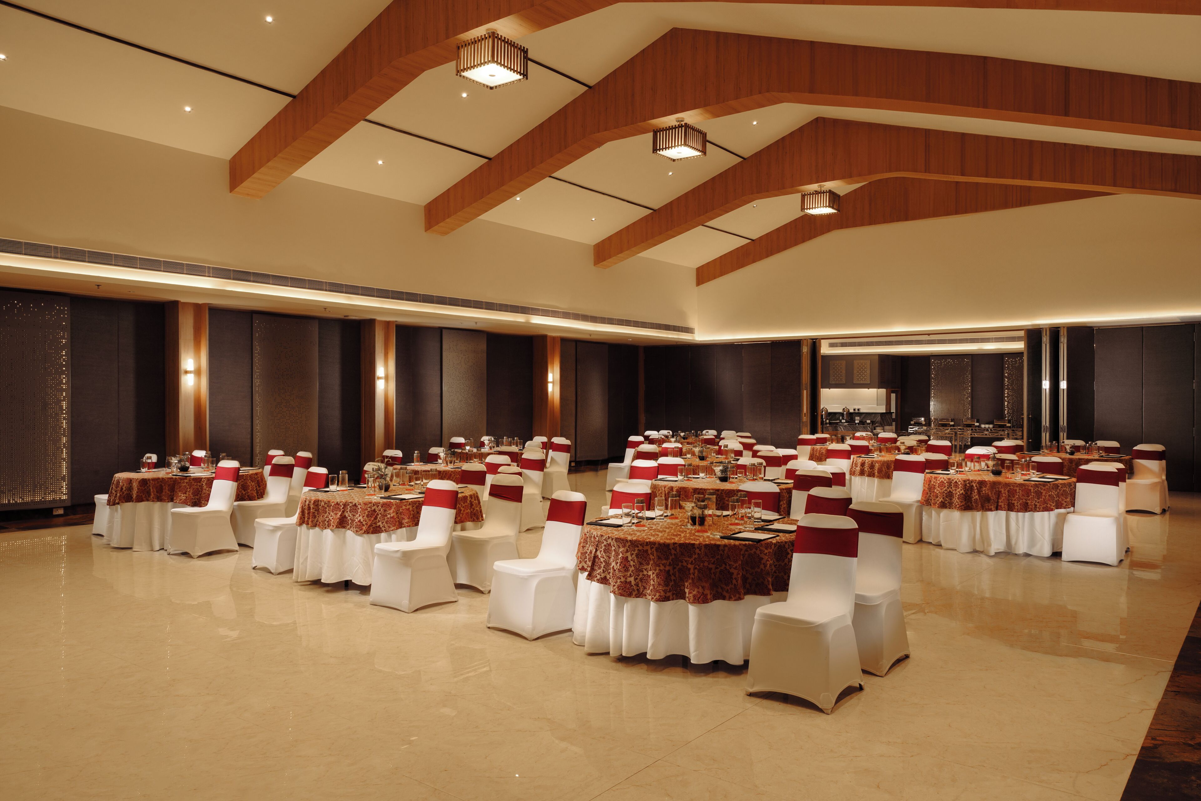 Un amplio salón de banquetes con varias mesas redondas, cada una rodeada de sillas blancas adornadas con cintas rojas. La sala cuenta con techos altos con vigas de madera e iluminación empotrada.