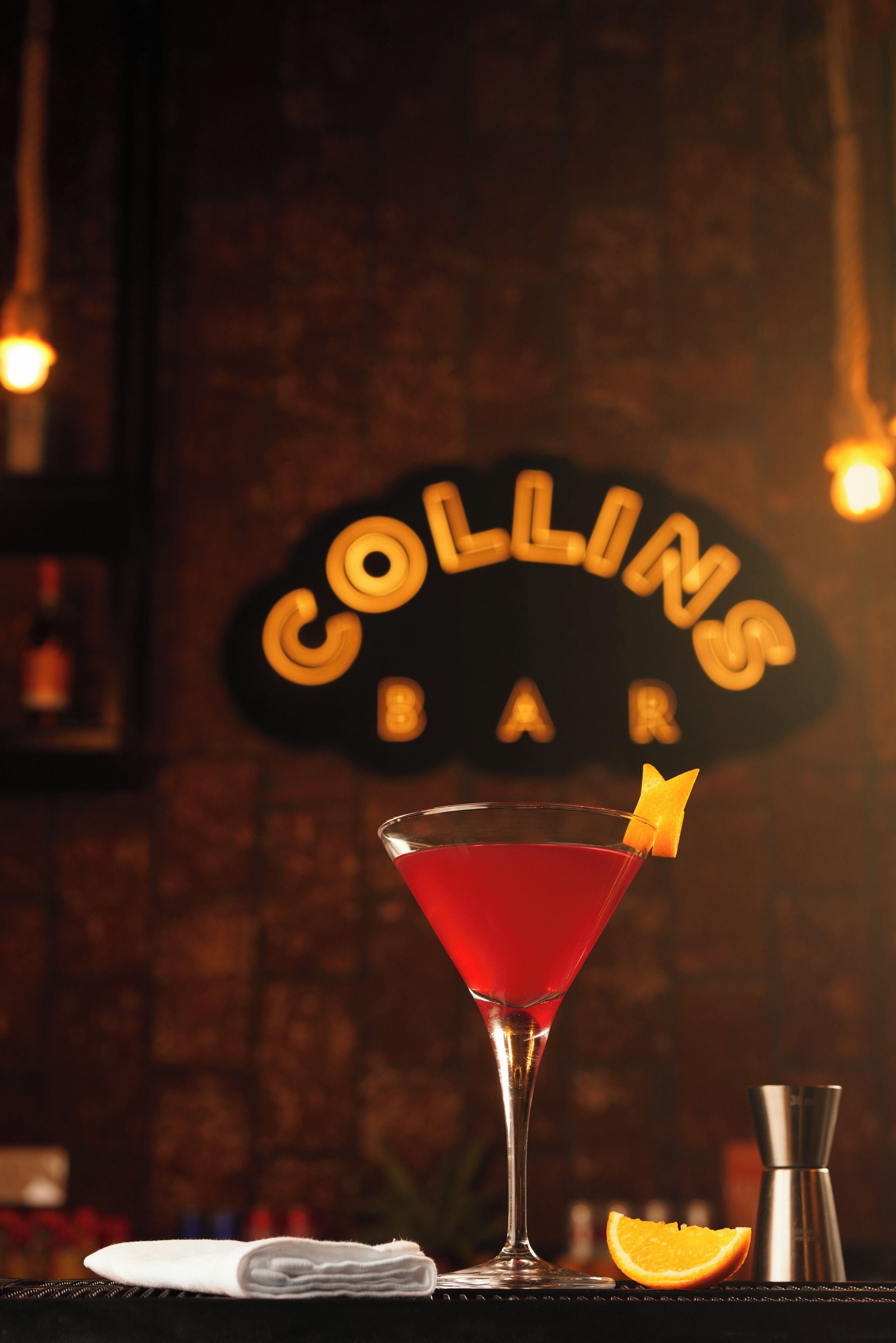A cocktail in a martini glass with an orange garnish, a cocktail shaker, and an orange slice on a bar counter.