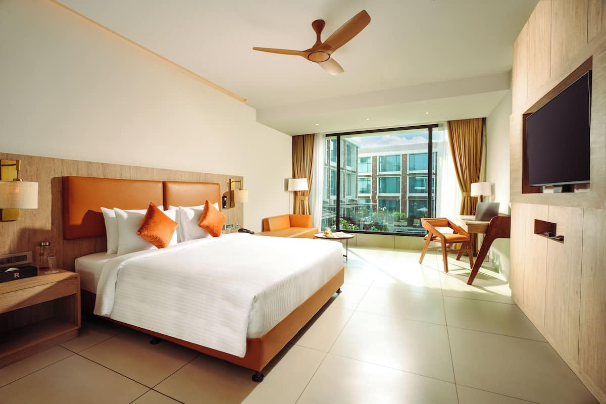 A modern hotel room with a large bed, a desk, a chair, a TV, and a balcony with a view.