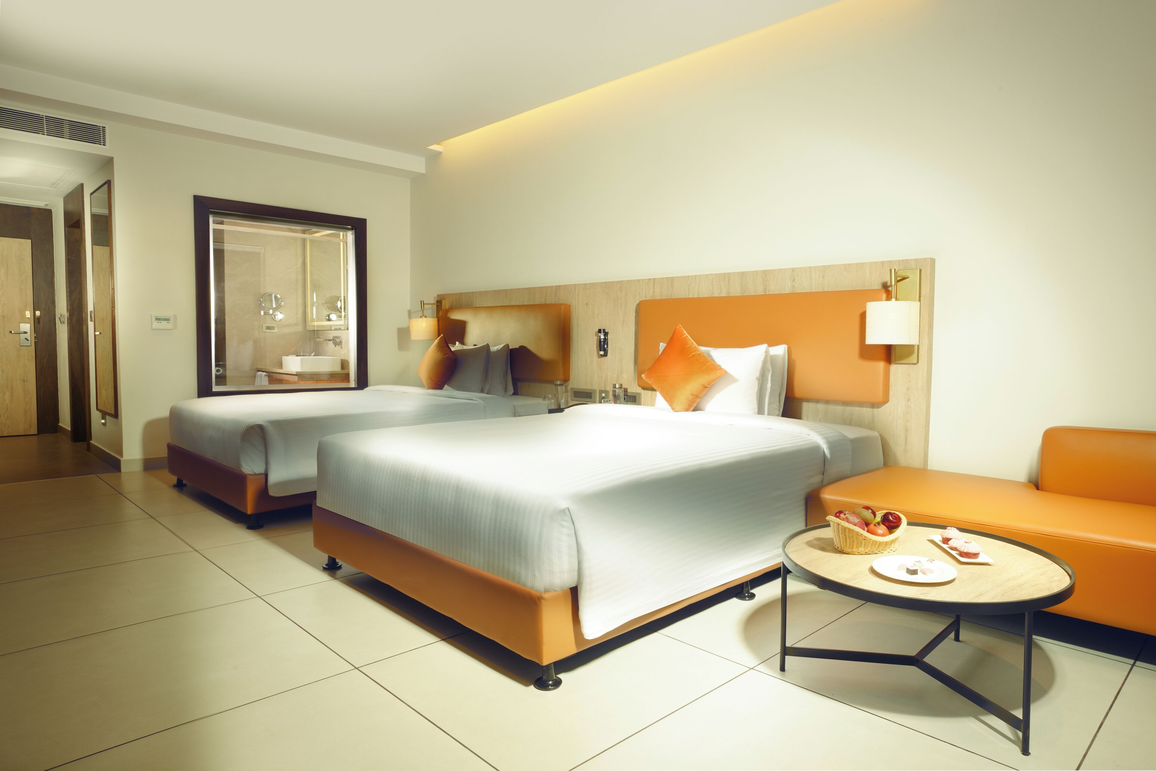 Quarto de hotel com duas camas, um sofá, uma mesa redonda com cesta e pratos, e um espelho grande.