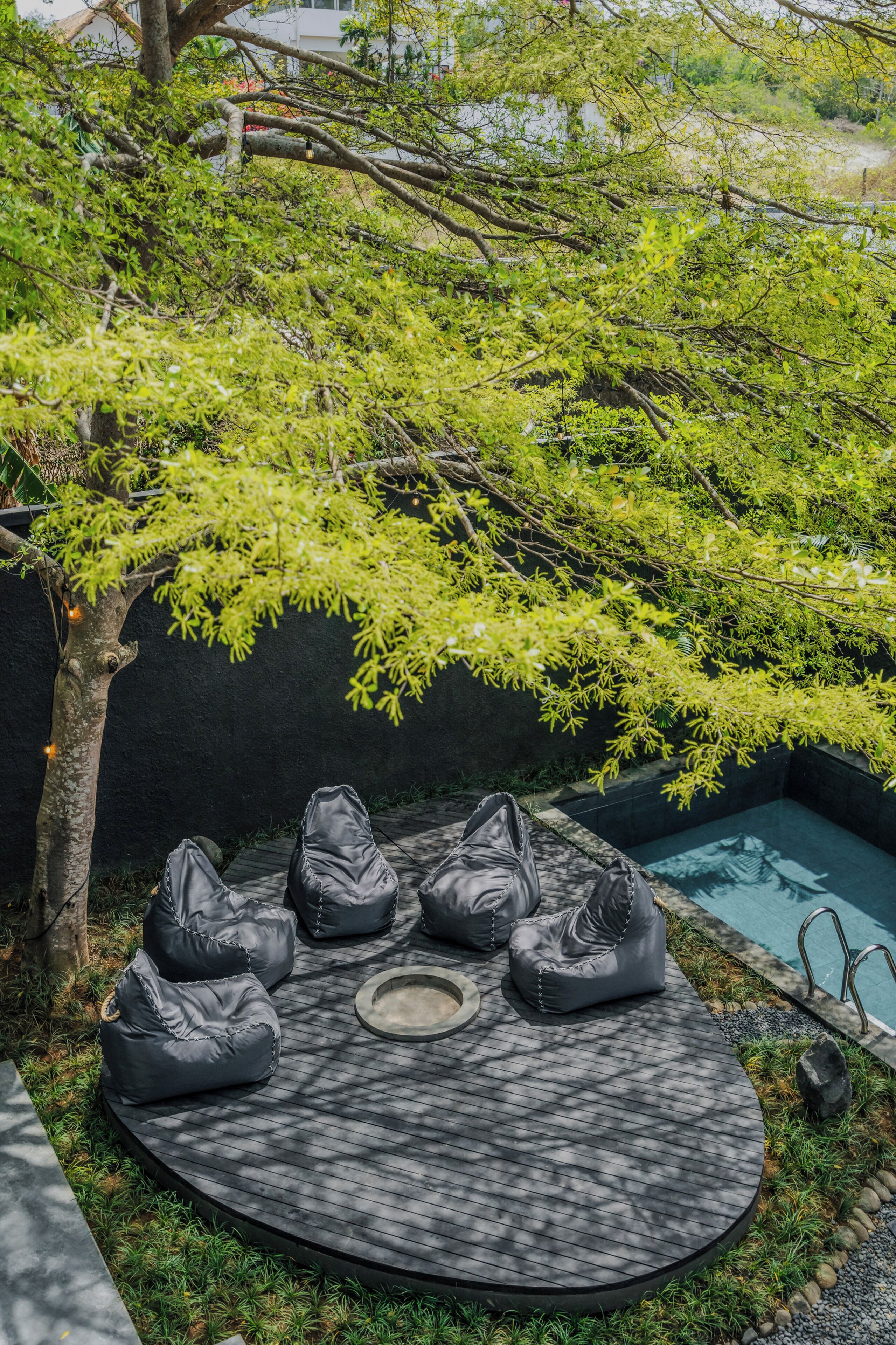 Área de estar circular com poltronas de fibra preta e uma mesa central, cercada por uma piscina e vegetação.