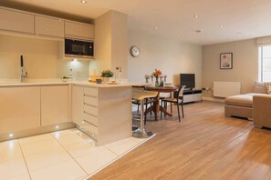 A modern kitchen with a microwave, a dining area with chairs, a sofa, and a clock on the wall. - Gated Parking! Quiet block, lift,  Sleeps up to 4 (Cheshire West and Chester)