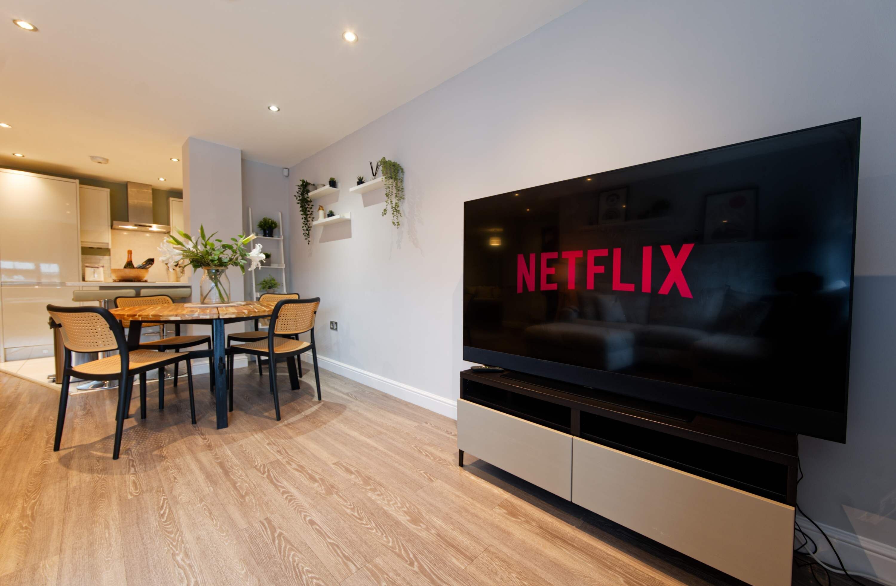 Una sala moderna con un televisor de pantalla plana grande montado en la pared, un comedor con mesa y sillas de madera, y una cocina al fondo.