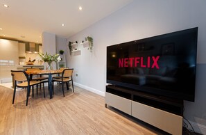 A modern living room with a large flat-screen TV mounted on the wall, a dining area with a wooden table and chairs, and a kitchen in the background. - Chester Apartment. Gated parking + Lift Sleeps up to 4 (Cheshire West and Chester)