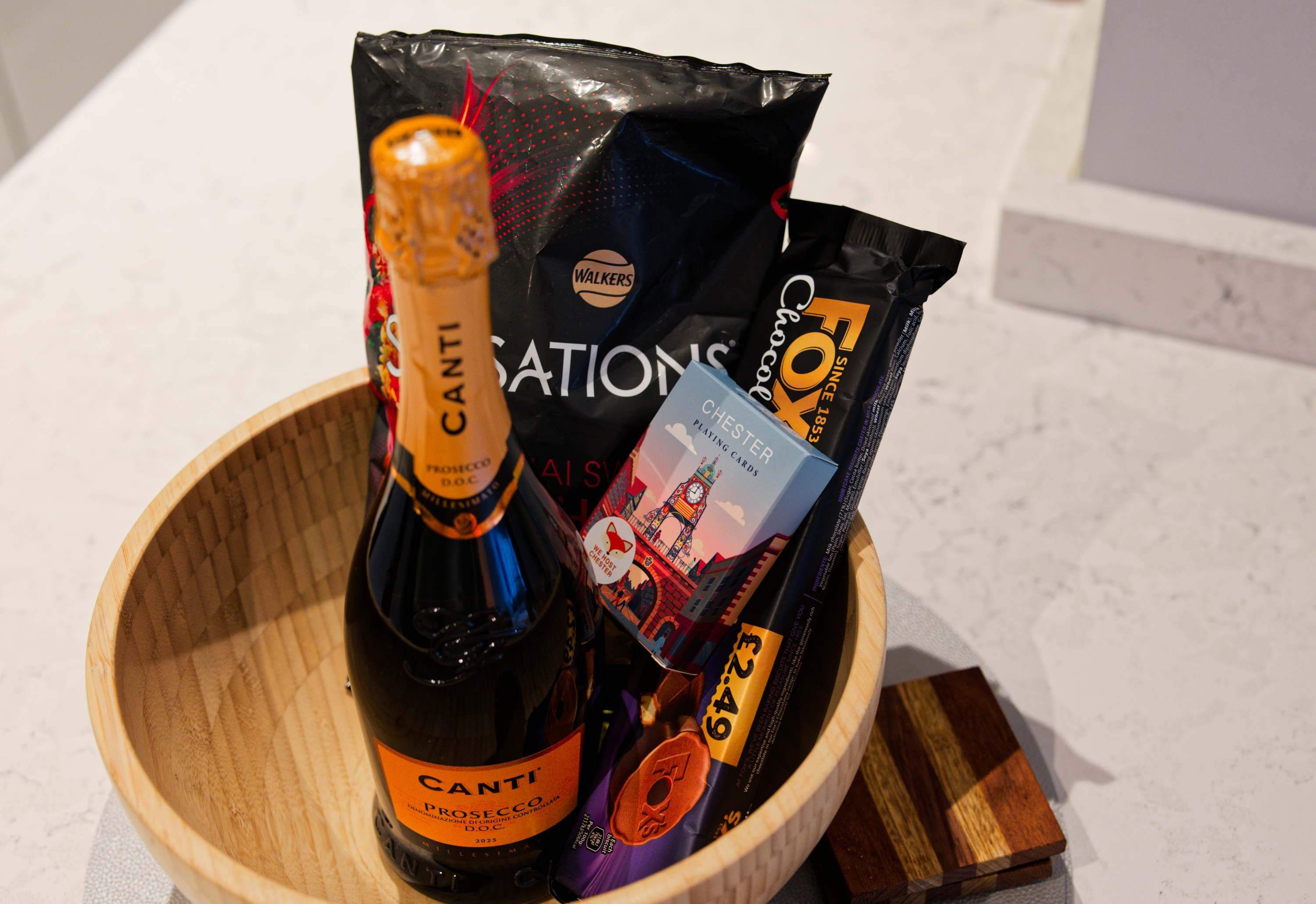 Un cuenco de madera que contiene una botella de Canti Prosecco, una bolsa negra con el logo de Fox y una caja azul de naipes Chester.