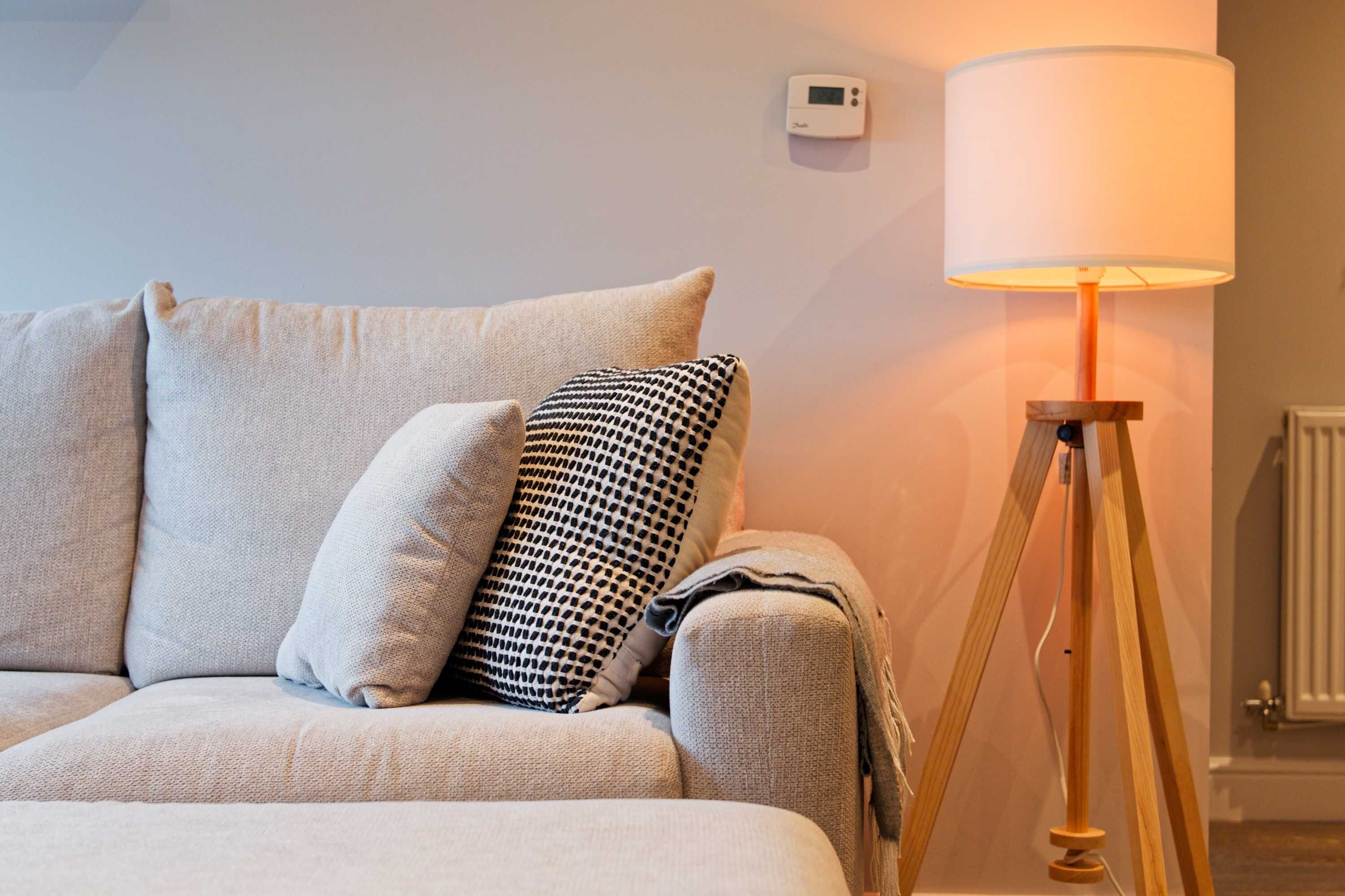 A room with a sofa, pillows, a lamp, and a thermostat on the wall.