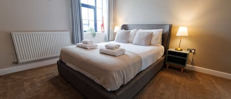A neatly made bed with a grey headboard, a bedside table with a lamp, and a radiator on the wall.