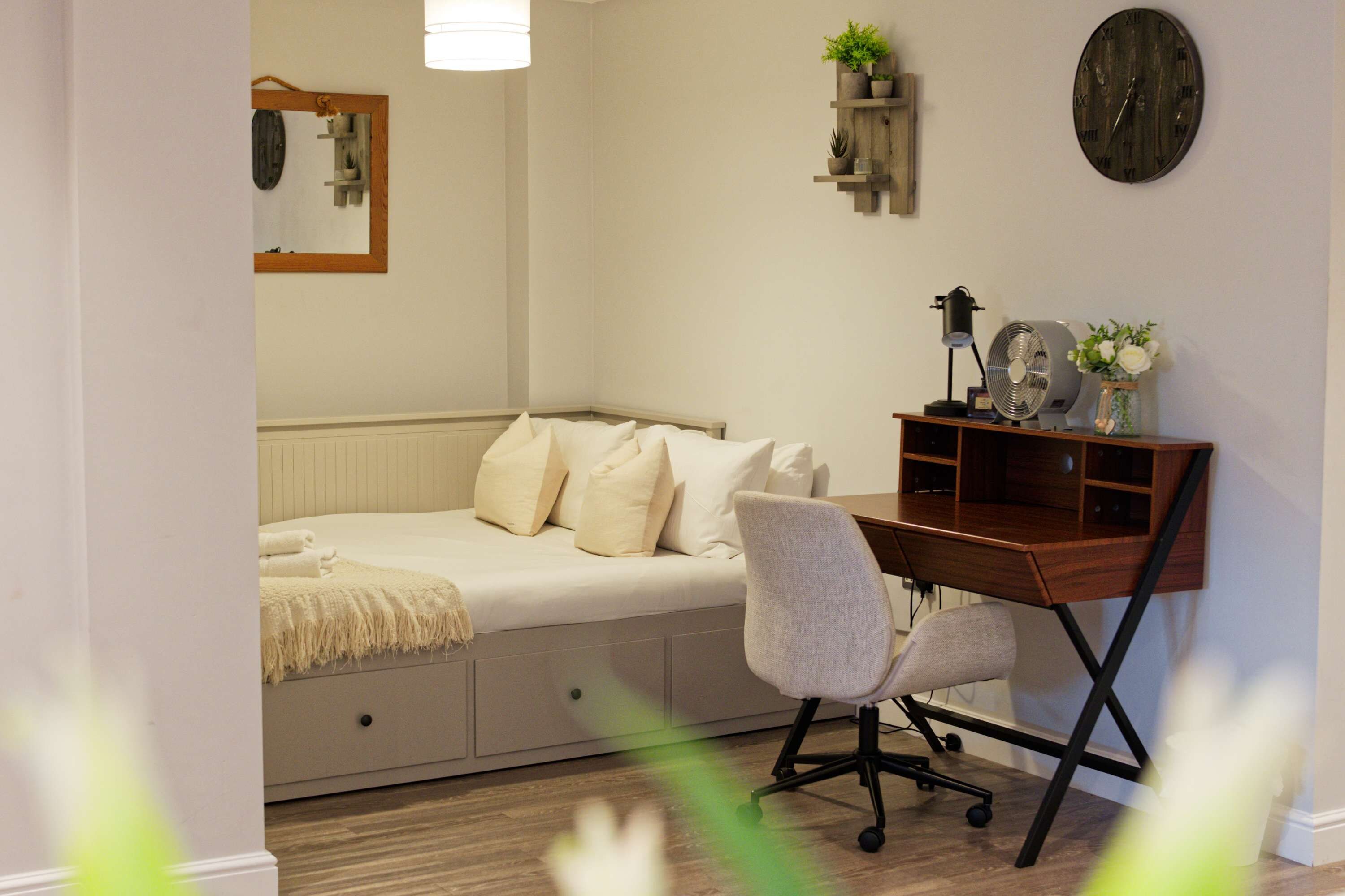 A bedroom with a bed, a desk, a chair, a clock, and a shelf with plants.