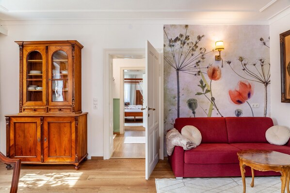 A room with a wooden cabinet, a red sofa, a wall with floral artwork, and a hallway leading to another room.