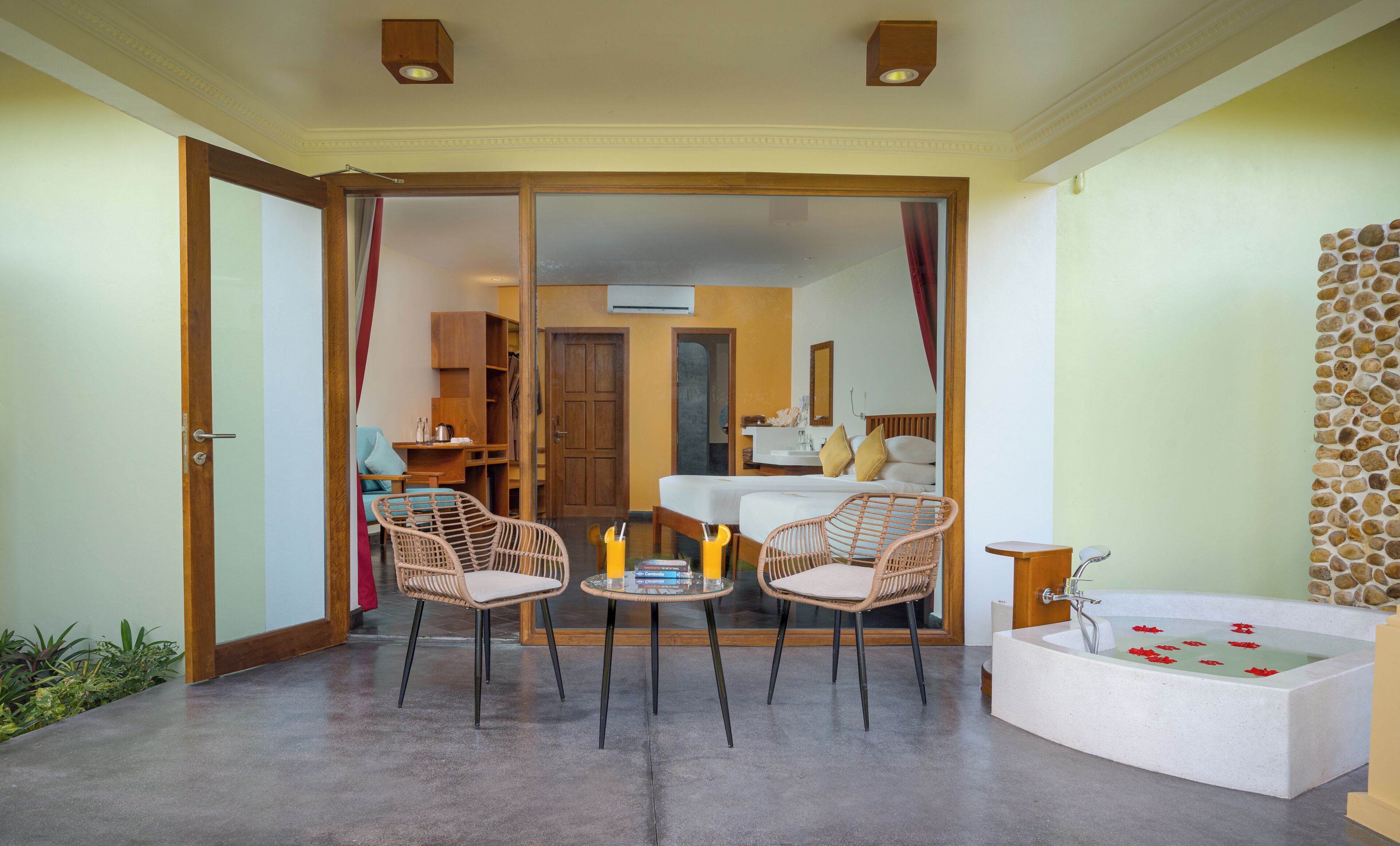A hotel room with a glass door leading to a balcony, a bathtub with rose petals, wicker chairs, a small table with books, and a wooden cabinet.