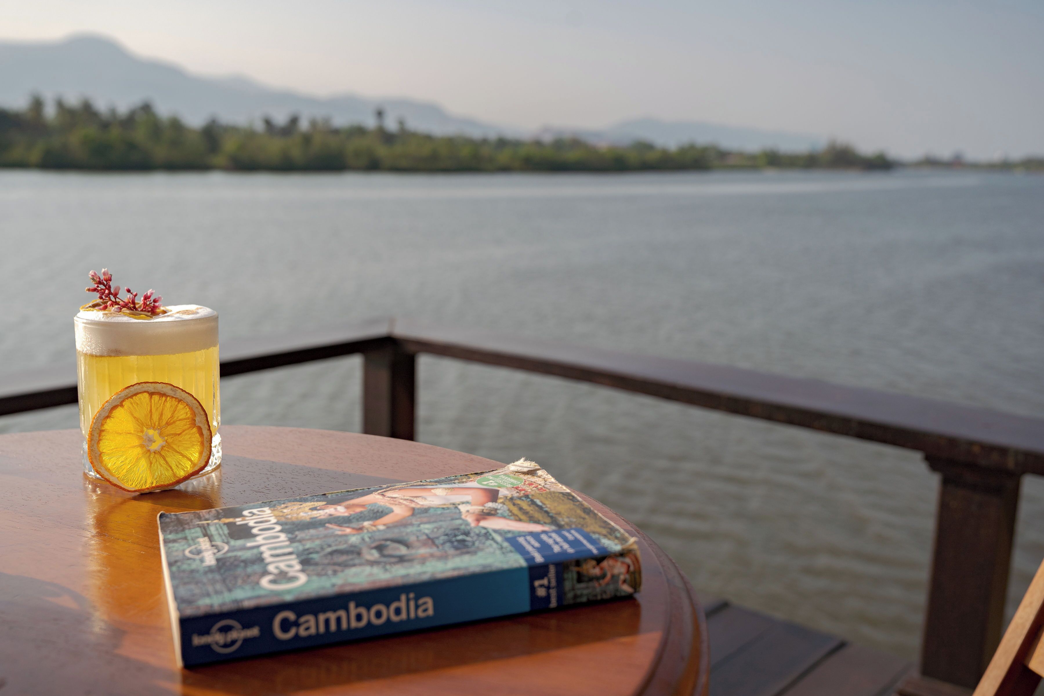 Una mesa con un libro titulado 'Cambodia' y una taza de té adornada con una rodaja de naranja deshidratada.