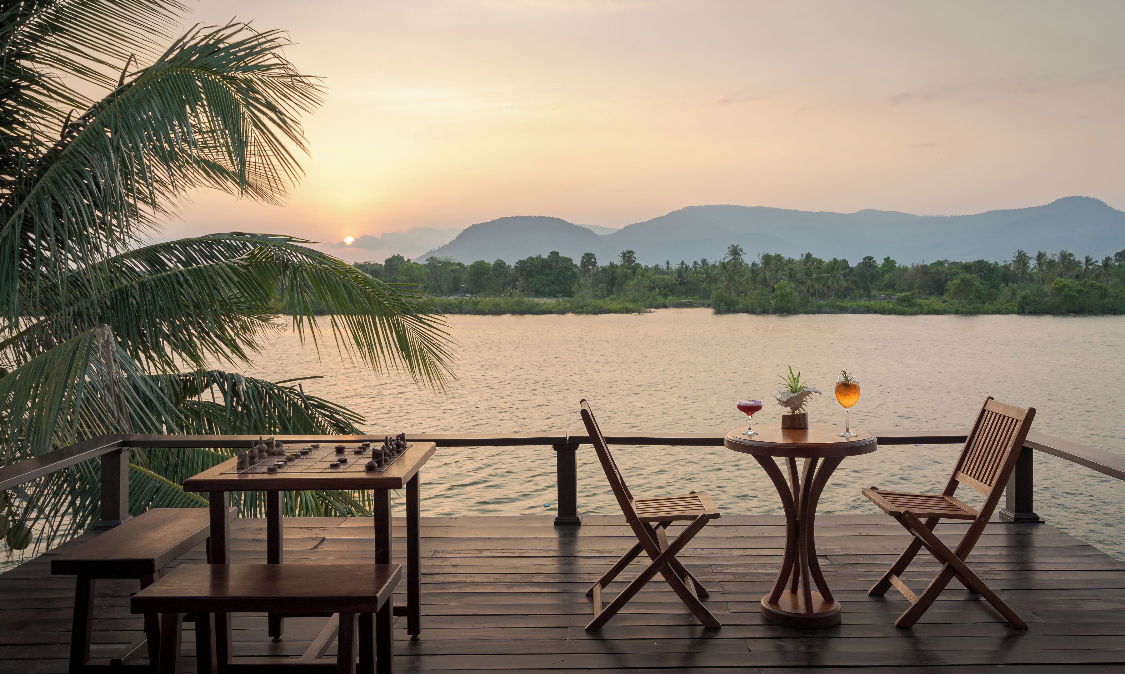 Une terrasse en bois, agrémentée de tables et de chaises, qui offre une vue sur un lac paisible, entouré de palmiers et de collines lointaines.