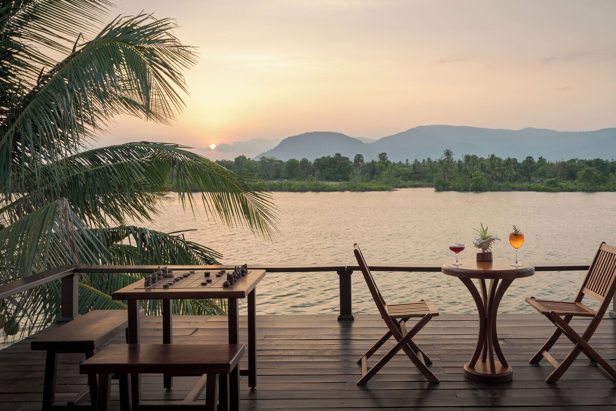 Une terrasse en bois, agrémentée de tables et de chaises, qui offre une vue sur un lac paisible, entouré de palmiers et de collines lointaines.