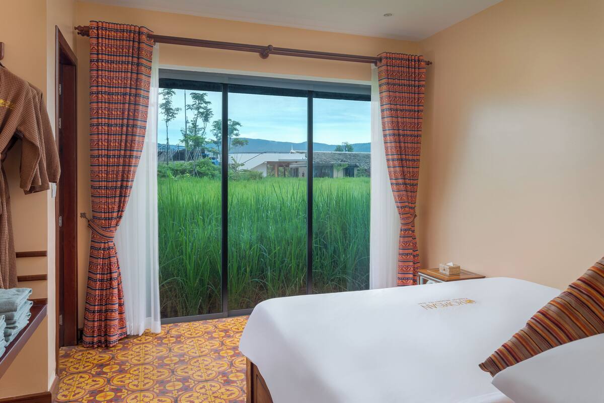 Ein Hotelzimmer mit einem großen Bett, Blick auf ein grünes Feld durch eine Schiebetür und einem kleinen Tisch mit einer Lampe.