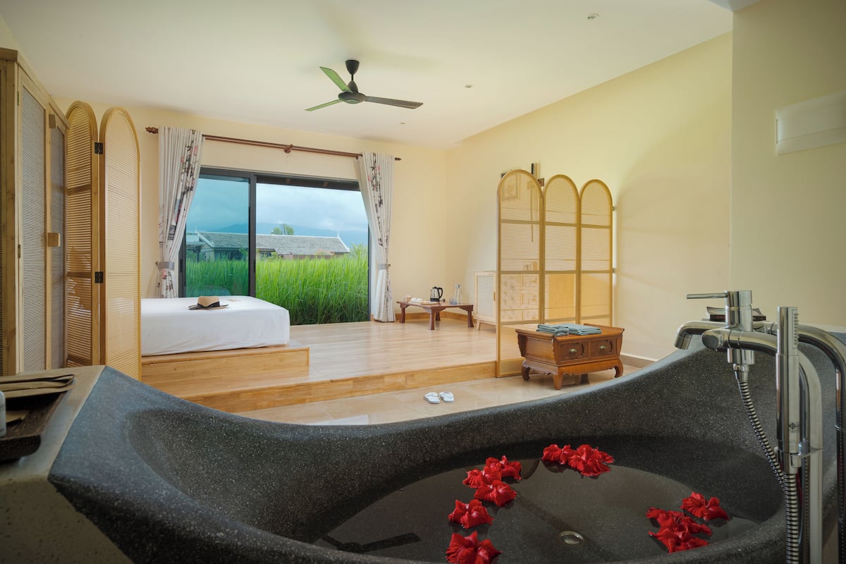 Ein Zimmer mit einer großen Badewanne, einem Bett, einem Holztisch und Blick auf ein grünes Feld durch ein Fenster.