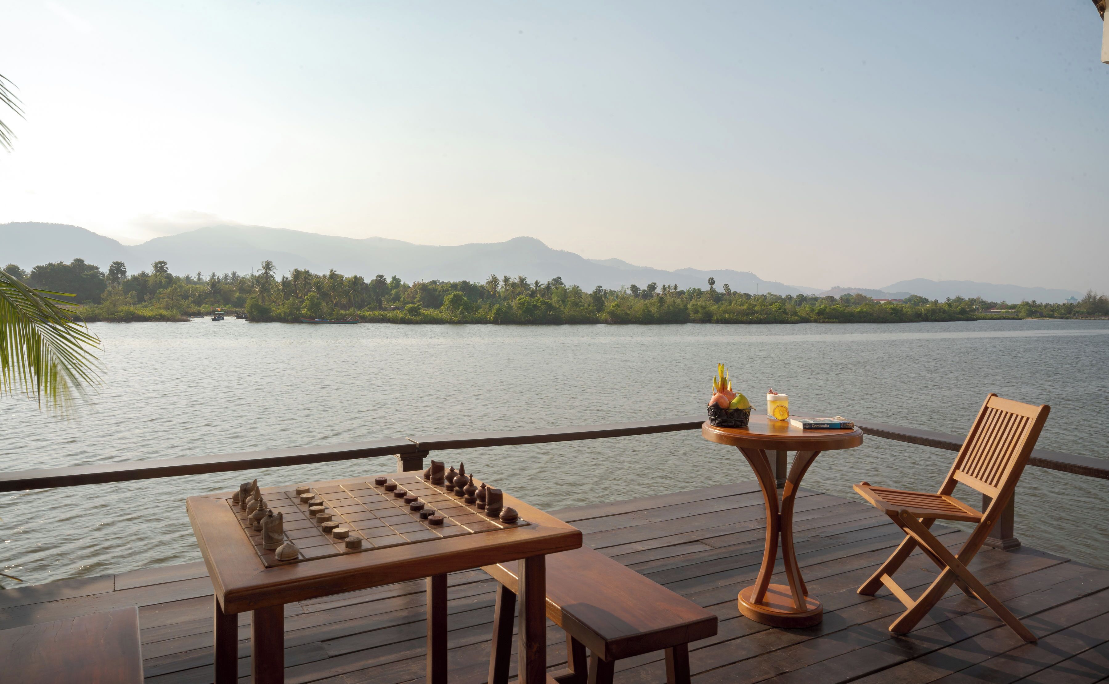A wooden deck with a chessboard, a table with a drink and a book, and a chair.
