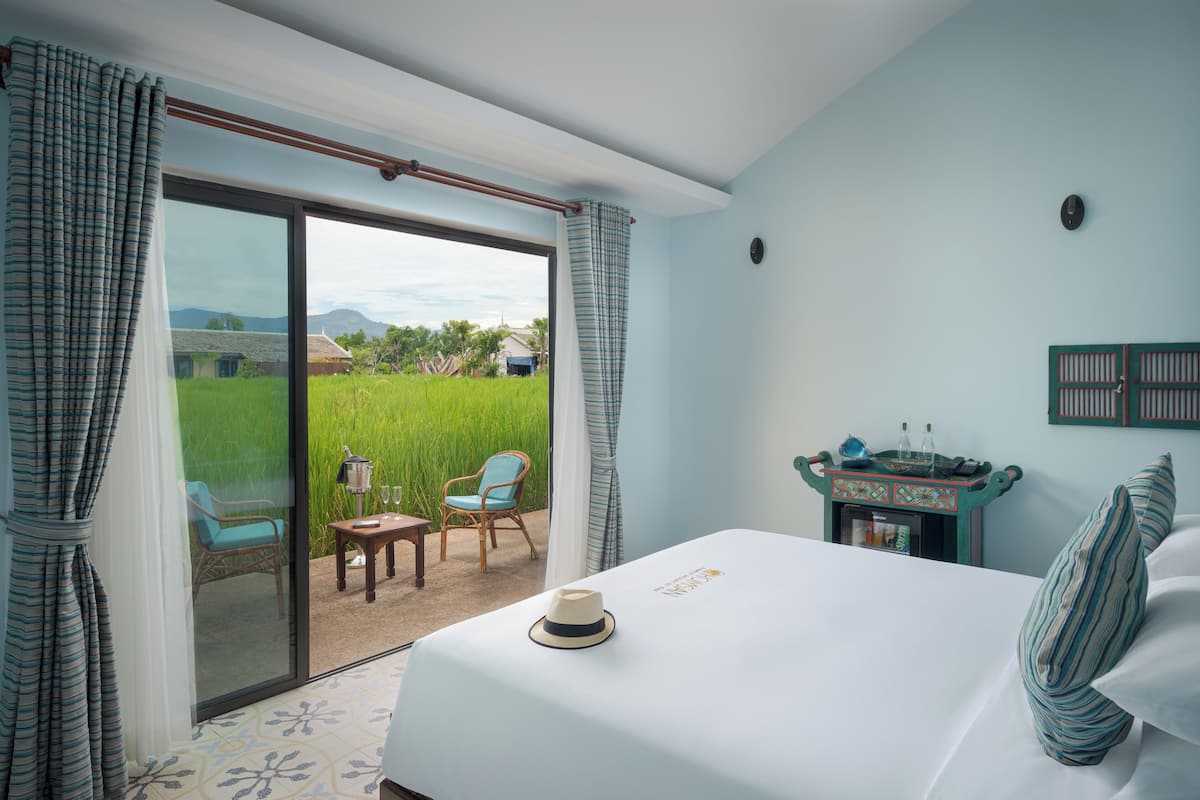 Ein Schlafzimmer mit einem großen Bett, Blick auf ein grünes Feld, einem kleinen Tisch mit Stuhl und einem Nachttisch mit Lampe.