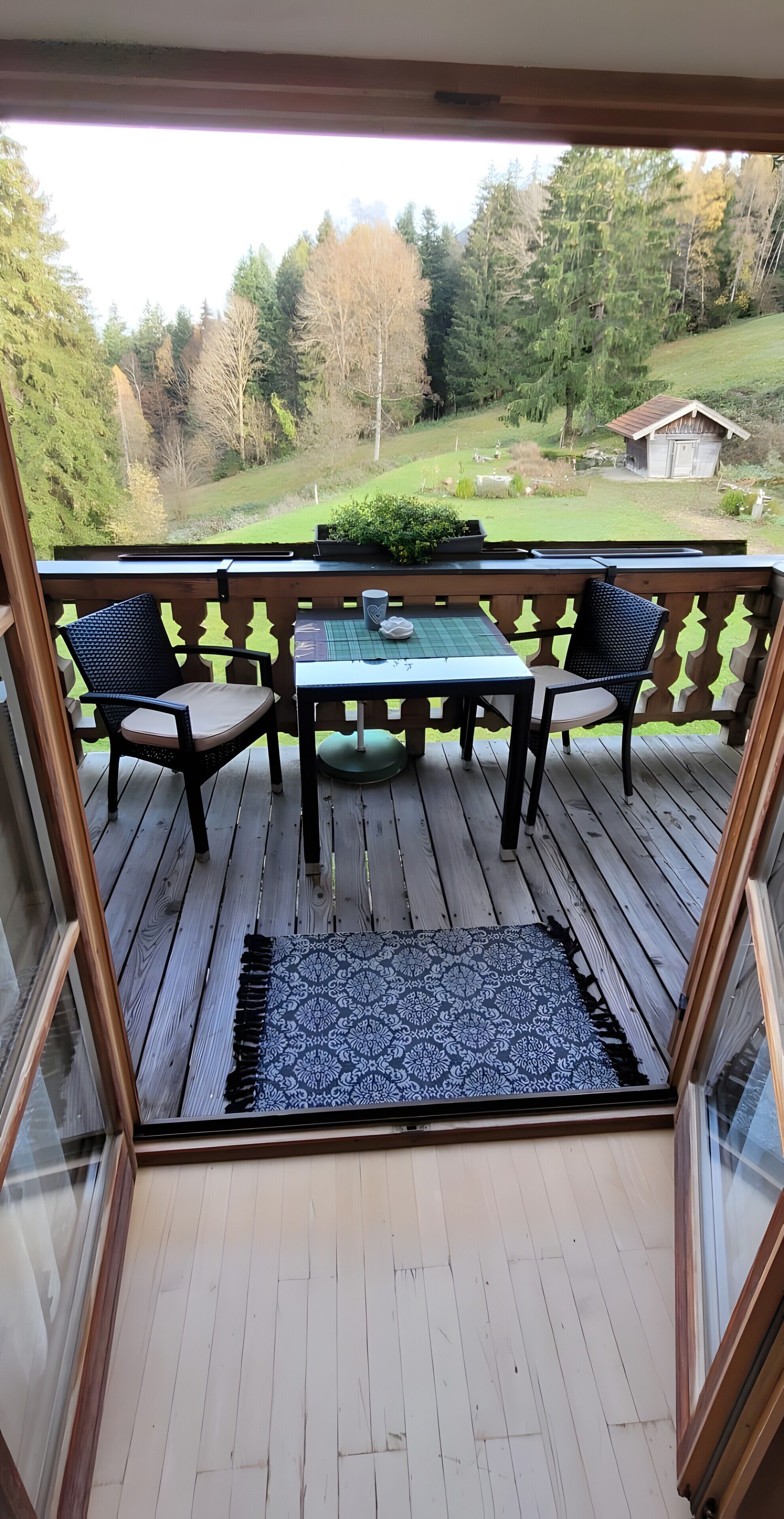 Een houten terras met een tafel en stoelen, een kamerbreed tapijt met patroon, en uitzicht op een groen landschap met bomen en een klein gebouw.