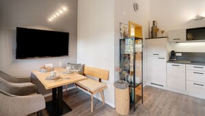 A modern kitchen with white cabinets, a dining area with a wooden table and bench, and a flat-screen TV mounted on the wall.