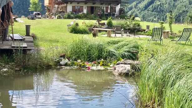 A chalet-style building with a balcony, surrounded by a lush garden and a small pond.