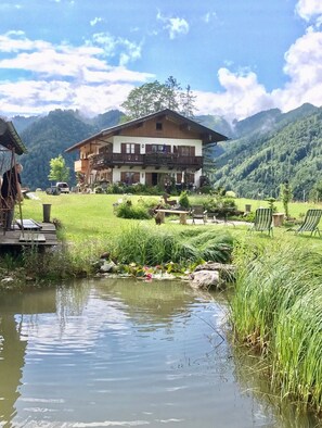 Un edificio de estilo chalet con balcón, rodeado de un jardín exuberante y un pequeño estanque.
