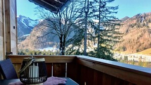 A wooden deck with a table and chairs, overlooking a mountainous landscape with trees and a building in the distance.