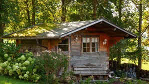 A rustic wooden cabin with a sloped roof, surrounded by lush greenery and trees.