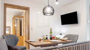 A modern dining area with a wooden table, two grey chairs, a wine bottle, glasses, and a TV mounted on the wall.