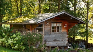 A rustic wooden cabin with a sloped roof, surrounded by lush greenery and trees.