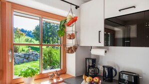 A kitchen with a large window, a hanging basket with vegetables, a coffee maker, a toaster, and a view of a green landscape. - Apartment 'Wiesengrund' with Garden View, Private Terrace and Shared Garden (Dietmannsried)