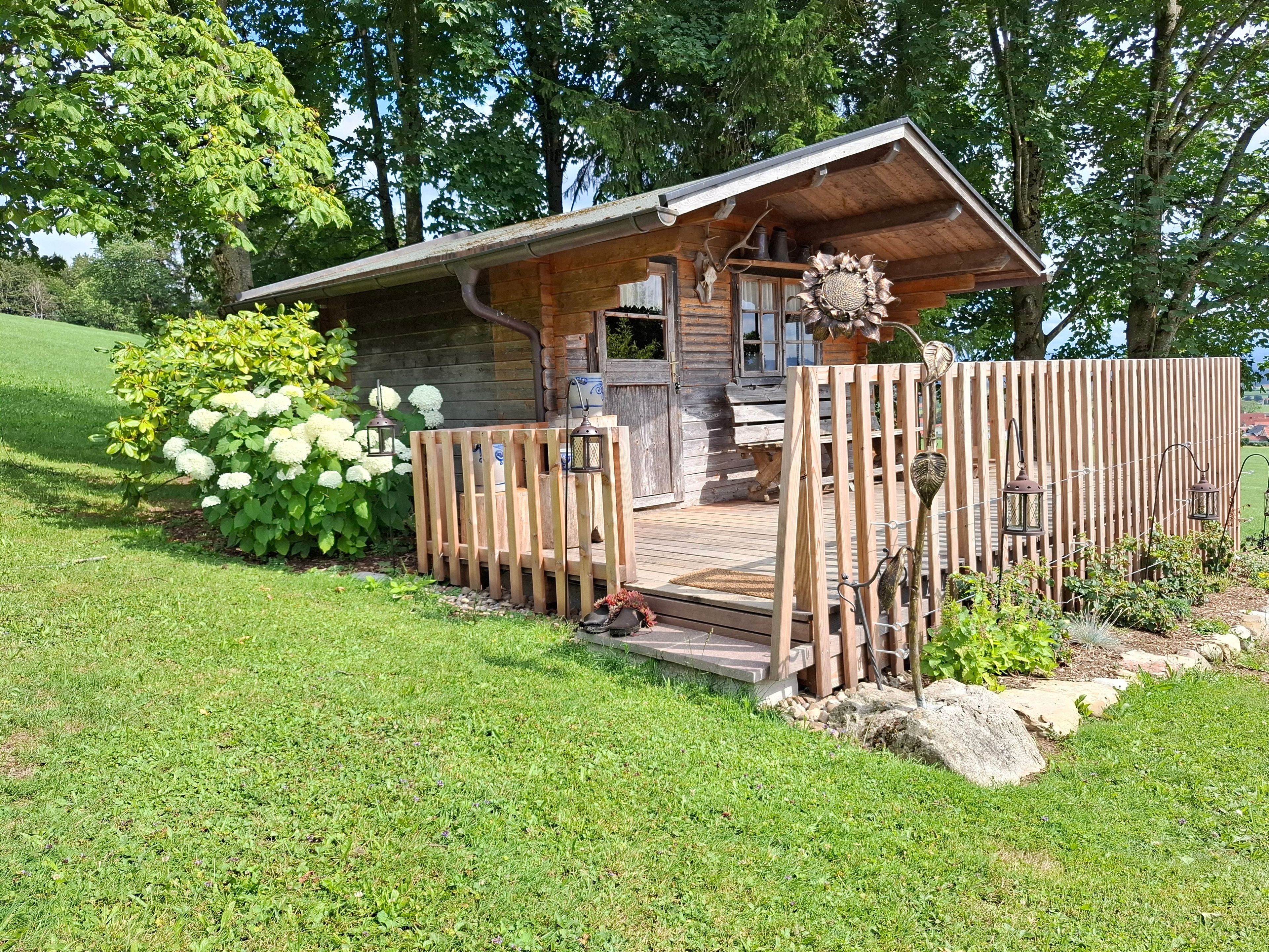 A rustic cabin with a wooden deck, surrounded by lush greenery and a wooden fence.