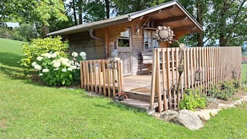 A rustic cabin with a wooden deck, surrounded by lush greenery and a wooden fence.